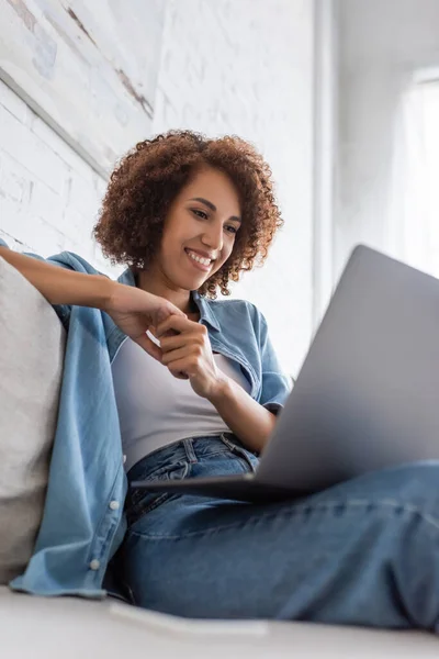 Vista de ángulo bajo de la mujer afroamericana feliz usando el ordenador portátil mientras está sentado en el sofá - foto de stock