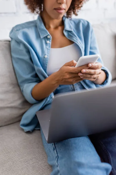 Обрізаний вид на афроамериканську жінку, що обмінюється повідомленнями на смартфоні біля ноутбука, сидячи на дивані — стокове фото