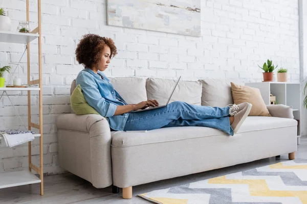 Focused african american woman using laptop while sitting on couch and working from home — Stock Photo