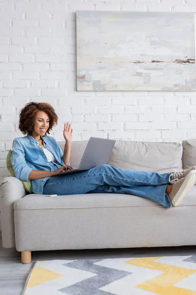 Feliz Africano americano mulher acenando mão durante vídeo gato no laptop enquanto sentado no sofá — Fotografia de Stock