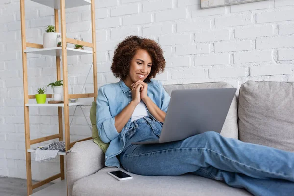 Mujer afroamericana feliz sentado con las manos apretadas y mirando el ordenador portátil cerca del teléfono inteligente en el sofá - foto de stock