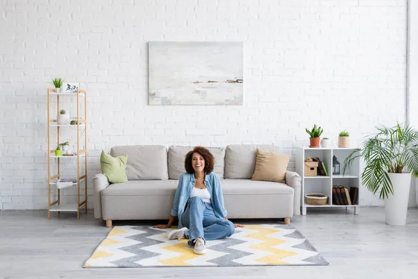 Femme afro-américaine gaie assise sur un tapis avec motif près du canapé moderne dans le salon — Photo de stock