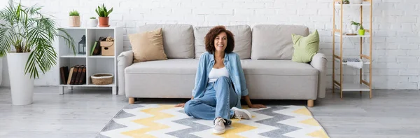Alegre mujer afroamericana sentada en la alfombra con patrón cerca de sofá moderno en la sala de estar, pancarta - foto de stock