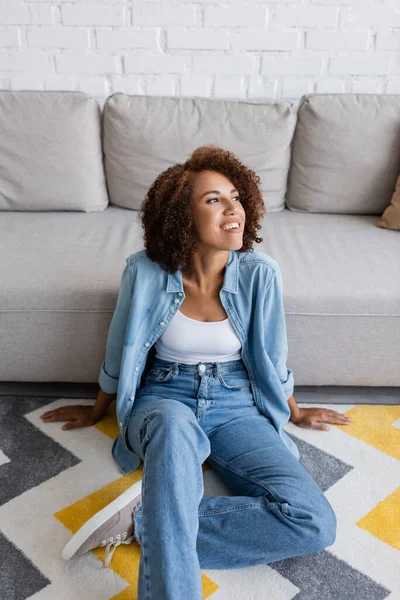 Sonriente mujer afroamericana sentada en la alfombra con el patrón cerca de sofá moderno en la sala de estar - foto de stock