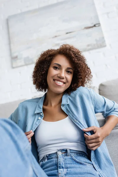 Mujer afroamericana sonriente sentada cerca de un sofá moderno en la sala de estar - foto de stock