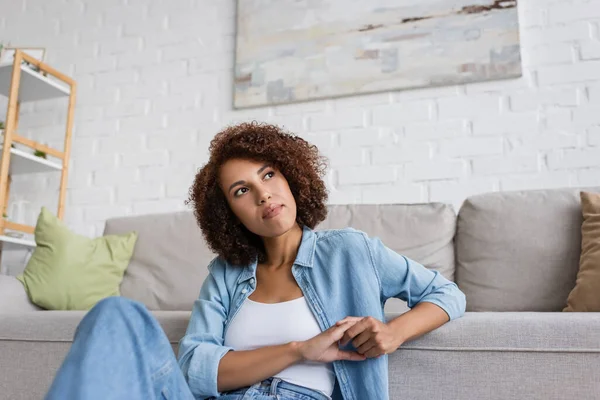 Donna afro-americana sognante seduta vicino al divano moderno in soggiorno — Foto stock