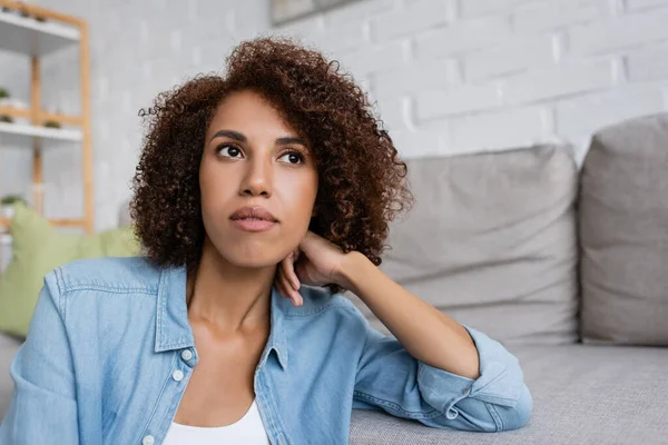 Стомлена афроамериканська жінка з кучерявим волоссям, що сидить біля сучасного дивана у вітальні — стокове фото