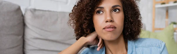 Pensive african american woman with curly hair sitting near modern sofa in living room, banner — Stock Photo