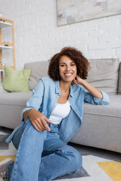 Lächelnde Afroamerikanerin mit lockigem Haar sitzt neben modernem Sofa im Wohnzimmer — Stockfoto