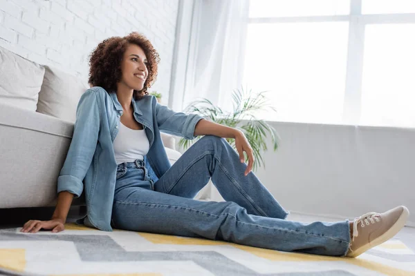 Задумчивая африканская американка в джинсовой одежде сидит рядом с современным диваном в гостиной — стоковое фото