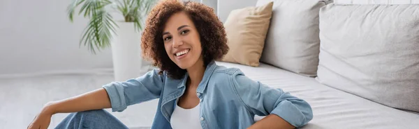 Позитивна афроамериканка з кучерявим волоссям, що сидить біля сучасного дивана у вітальні, банер — стокове фото