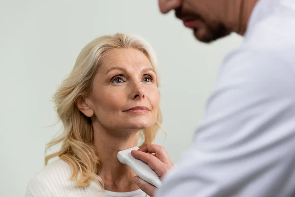 Mujer rubia mirando al doctor borroso examinando su garganta con ultrasonido - foto de stock