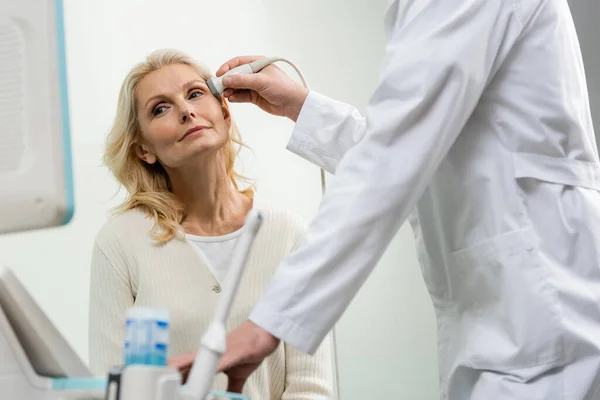 Mujer rubia mirando a la máquina de ultrasonido mientras el médico examina su cabeza - foto de stock