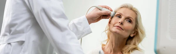 Mujer rubia mirando hacia otro lado cerca del médico haciendo ultrasonido examen de su cabeza, pancarta - foto de stock