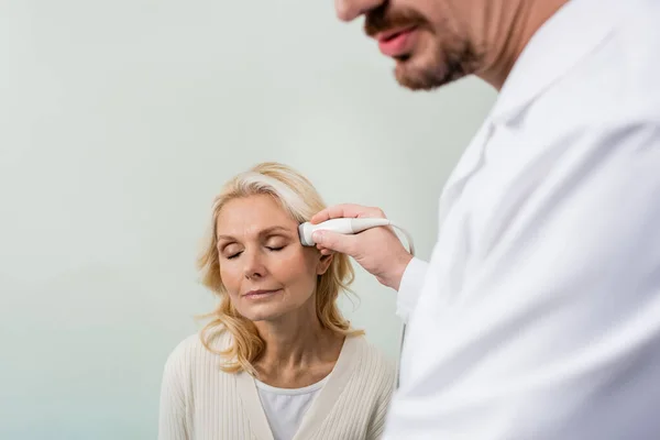 Médico desfocado fazendo ultra-som cabeça de mulher loira sentada com os olhos fechados — Fotografia de Stock