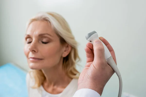 Medico che tiene sonda a ultrasuoni vicino alla donna bionda con gli occhi chiusi su sfondo sfocato — Foto stock