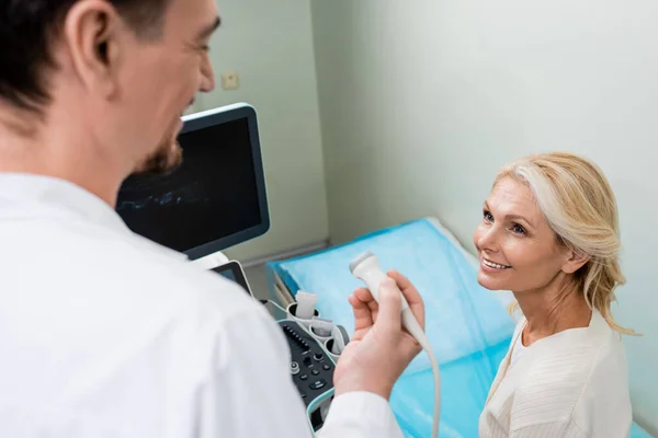 Doctor borroso sosteniendo sonda de ultrasonido cerca de mujer rubia positiva en la clínica - foto de stock