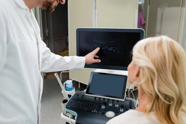 Médico apontando para a imagem no monitor de ultra-som perto de mulher loira em primeiro plano borrado — Fotografia de Stock