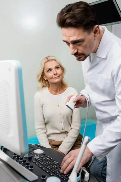 Внимательный врач смотрит на монитор ультразвукового аппарата рядом с улыбающейся женщиной средних лет — стоковое фото