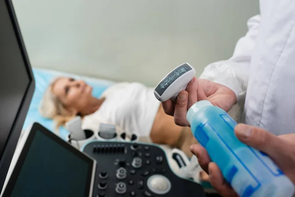 Medico che applica gel ultrasonico sulla sonda ad ultrasuoni vicino al paziente sdraiato su sfondo sfocato — Foto stock