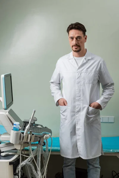 Médico morena de pie con las manos en bolsillos de capa blanca cerca de la máquina de ultrasonido - foto de stock