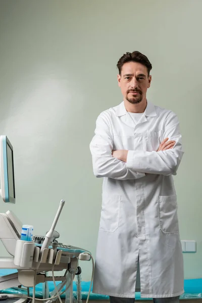 Serious doctor standing with crossed arms near ultrasound machine and looking at camera — Stock Photo