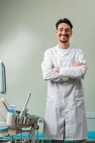 Médecin joyeux debout avec les bras croisés près de la machine ultrasonore moderne — Photo de stock