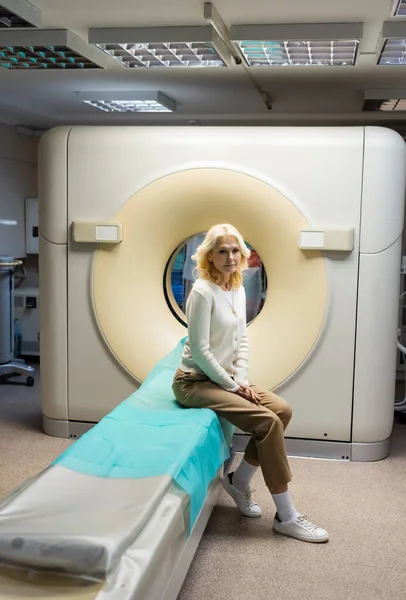 Comprimento total da mulher madura loira sentada perto da máquina de tomografia computadorizada na clínica — Fotografia de Stock