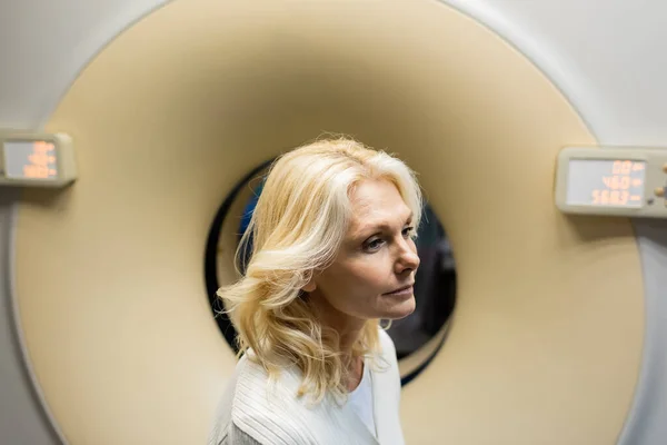 Rubia mujer de mediana edad mirando hacia otro lado cerca de la máquina de tomografía computarizada en el hospital - foto de stock