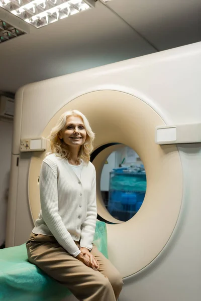 Веселая зрелая женщина сидит рядом с компьютерным томографом и смотрит в камеру — стоковое фото