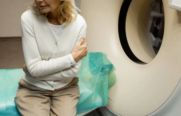 Vista cortada da mulher preocupada sentada com braços cruzados perto de tomógrafo na clínica — Fotografia de Stock