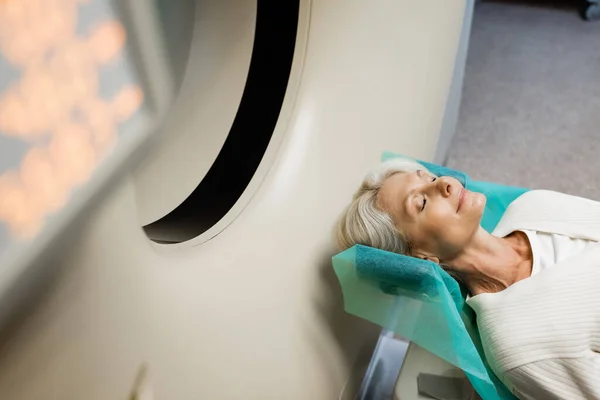 Vue grand angle de la femme mature avec les yeux fermés faire des diagnostics sur ct scanner sur le premier plan flou — Photo de stock