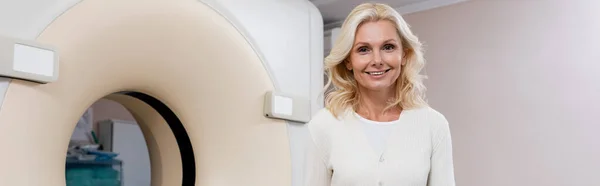 Glückliche blonde Frau lächelt in die Kamera in der Nähe des Scanners im Krankenhaus, Banner — Stockfoto