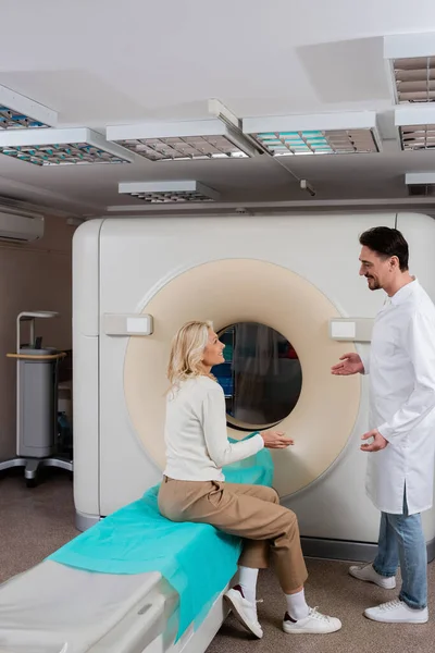 Вид сбоку положительного разговора врача и пациента и жестов рядом с компьютерным томографом в клинике — стоковое фото