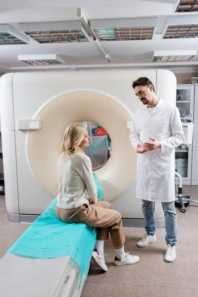 Longitud completa de radiólogo en bata blanca hablando con la mujer rubia sentada cerca de escáner ct en la clínica - foto de stock
