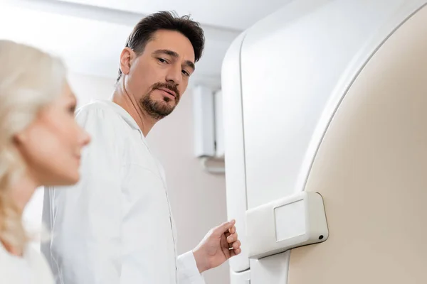 Radiologista grave olhando para mulher de meia-idade borrada perto do scanner de tomografia computadorizada no hospital — Fotografia de Stock