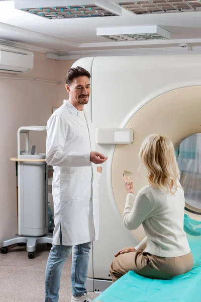 Позитивный доктор в белом халате разговаривает со зрелой женщиной, сидящей рядом с компьютерным томографом — стоковое фото