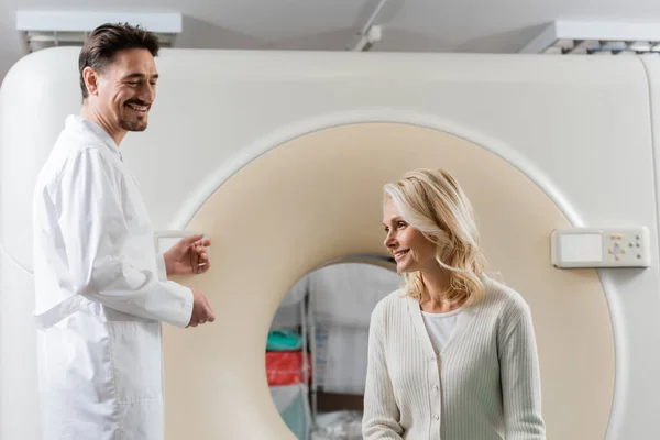 Fröhlicher Radiologe schaut lächelnde reife Frau in der Nähe von Computertomographen an — Stockfoto
