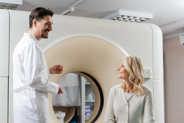 Blonde middle aged woman and positive radiologist looking at each other near ct scanner in clinic — Stock Photo