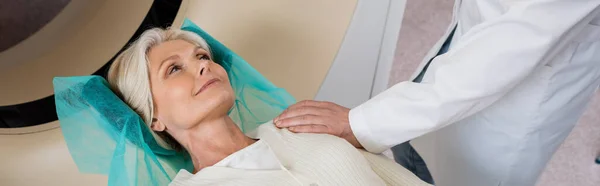 Рентгенолог трогает плечо положительной женщины среднего возраста перед компьютерной томографией в клинике, баннер — стоковое фото