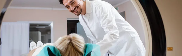 Radiologista em casaco branco sorrindo perto da mulher e máquina de tomografia computadorizada, banner — Fotografia de Stock