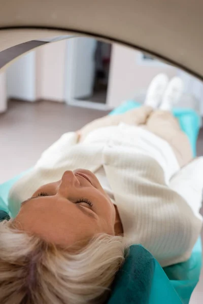 Femme mature avec les yeux fermés couché pendant les diagnostics sur ct scanner sur fond flou — Photo de stock