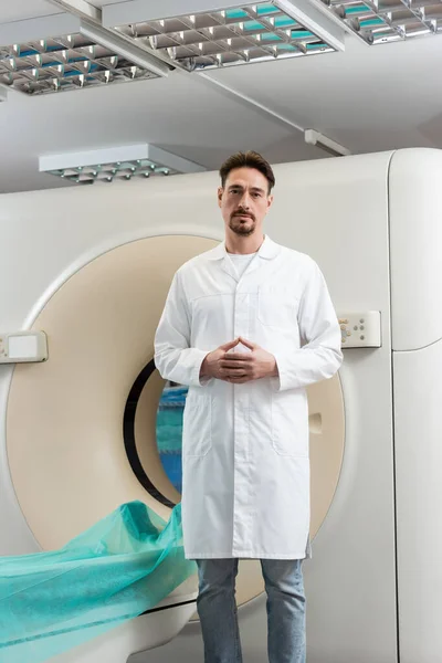 Schwerbärtiger Radiologe blickt in Klinik auf Kamera in der Nähe von Computertomographie-Scanner — Stockfoto