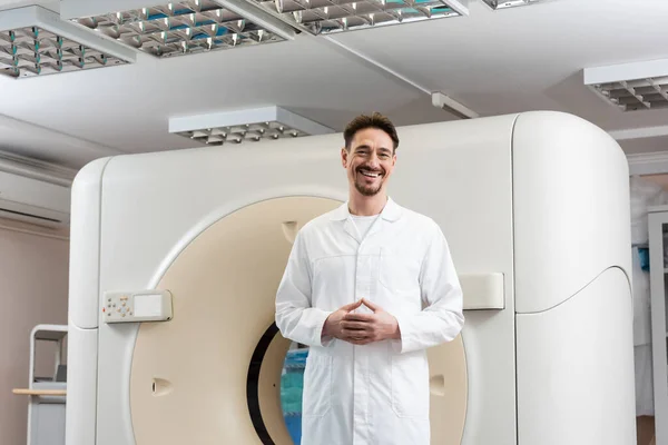 Щасливий радіолог у білому пальто, що стоїть біля комп'ютерного томографічного сканера і дивиться на камеру — стокове фото