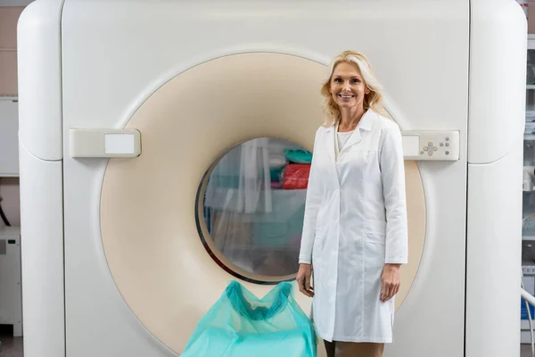 Hübsche blonde Radiologin im weißen Kittel lächelt in die Kamera neben dem Computertomographen — Stockfoto