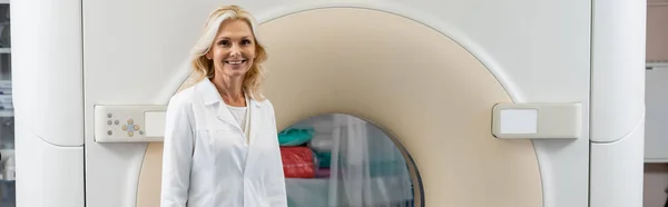 Усміхнена блондинка-радіолог дивиться на камеру біля комп'ютерної томографічної машини, банер — стокове фото