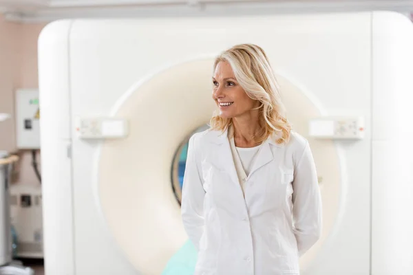Lächelnder blonder Arzt in weißem Mantel schaut in der Nähe des Scanners im Krankenhaus weg — Stockfoto
