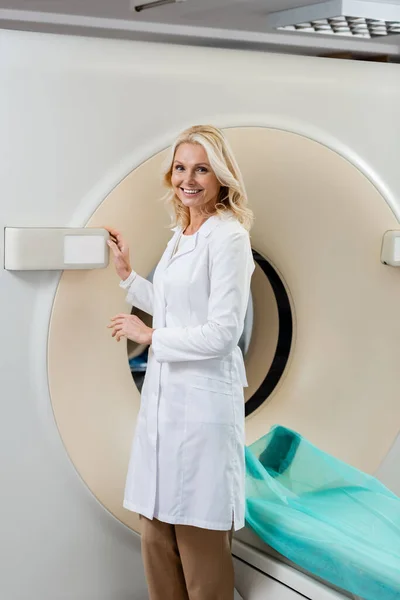 Blonder Radiologe im weißen Kittel lächelt in die Kamera neben Computertomographen — Stockfoto