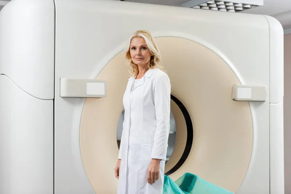 Radiologista de meia idade em casaco branco em pé perto tomografia computadorizada scanner e olhando para a câmera — Fotografia de Stock