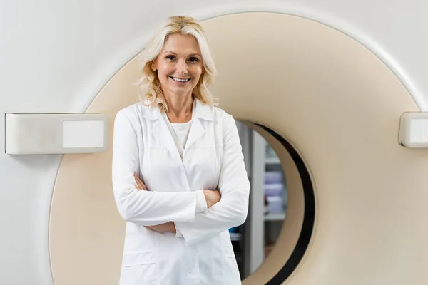 Joyful physician in white coat standing with crossed arms near computed tomography scanner — Stock Photo